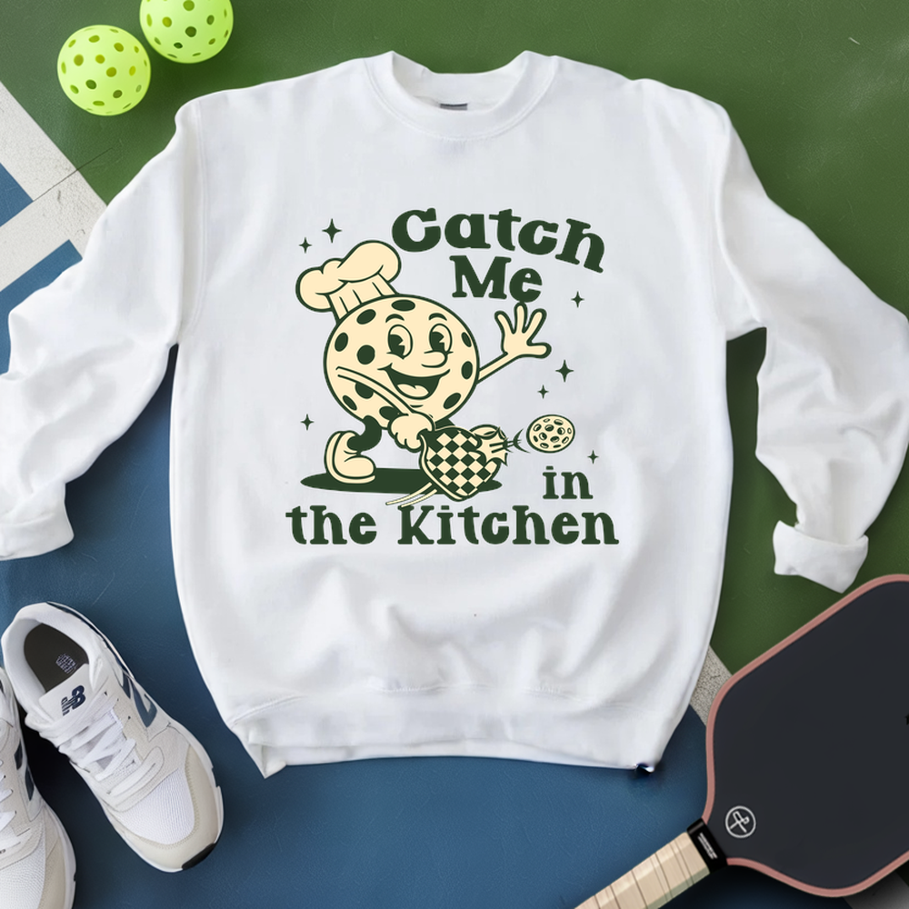 White sweatshirt with a cartoon character and text 'Catch Me in the Kitchen' on a green and blue surface with pickleball equipment.