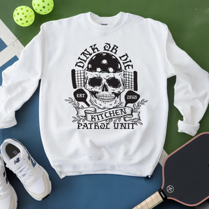White sweatshirt with skull design and text, placed on a green and blue surface with pickleball equipment.
