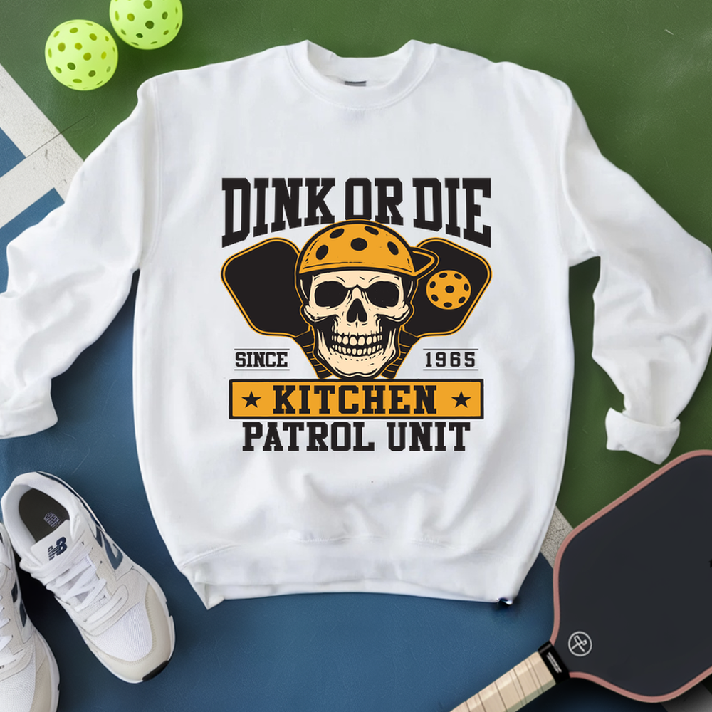 White sweatshirt with skull graphic and text on a green and blue surface with pickleballs and a paddle.
