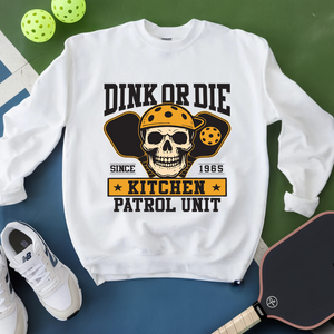 White sweatshirt with skull graphic and text on a green and blue surface with pickleballs and a paddle.