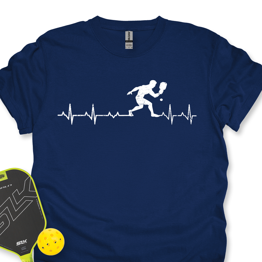 Navy blue t-shirt with pickleball player design and heart rate line, next to a paddle and ball on a white background.
