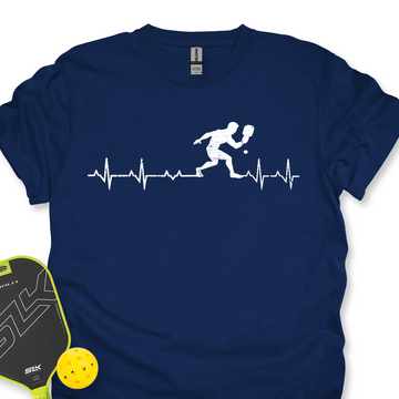 Navy blue t-shirt with pickleball player design and heart rate line, next to a paddle and ball on a white background.