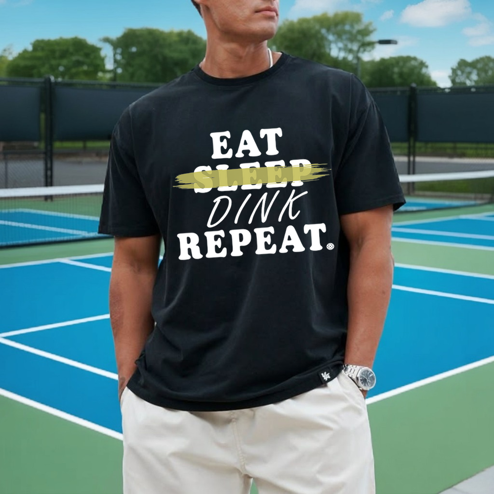 Person wearing a black t-shirt with 'EAT SLEEP DINK REPEAT' text on a tennis court.