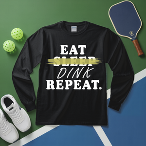 Black long-sleeve shirt with 'Eat Sleep Dink Repeat' text on a sports court background with pickleball equipment.