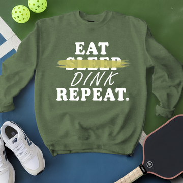 Green sweatshirt with 'Eat Sleep Dink Repeat' text, white sneakers, and pickleball paddles on a green and blue background