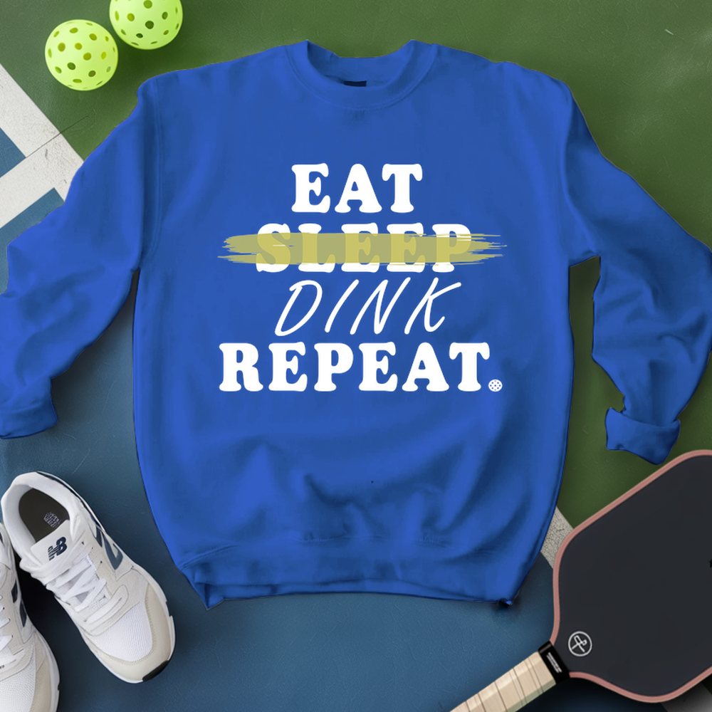 Blue sweatshirt with 'EAT SLEEP DINK REPEAT' text on a green surface with pickleball equipment.