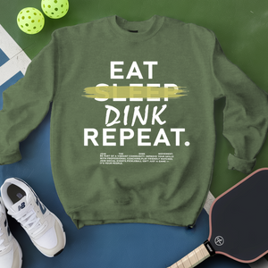 Green sweatshirt with 'Eat Sleep Dink Repeat' text, pickleball balls, paddle, and shoes on a green and blue background