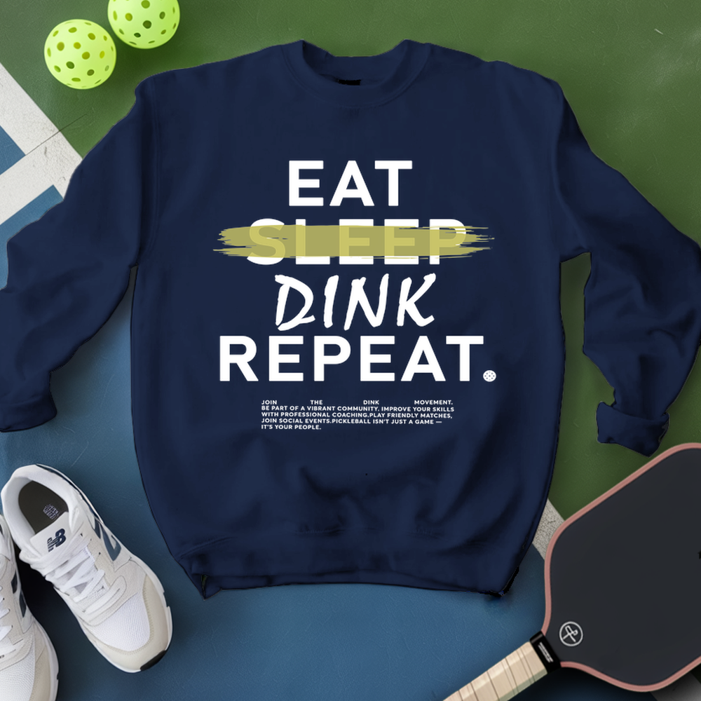 Navy blue sweatshirt with 'Eat Sleep Dink Repeat' text, next to a pickleball paddle and balls on a green surface.