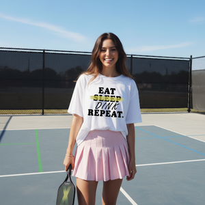 Woman on a tennis court wearing a white t-shirt with text and a pink skirt.