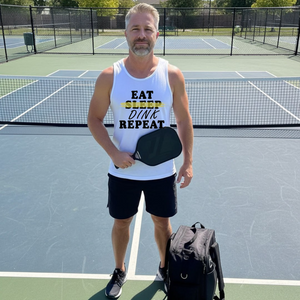 Man on a pickleball court wearing a tank top with text, holding a paddle and bag.