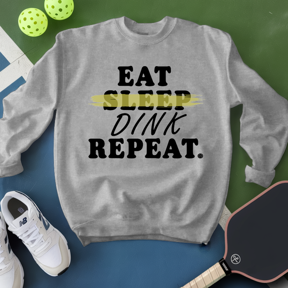Gray sweatshirt with 'EAT SLEEP DINK REPEAT' text, surrounded by pickleballs and a paddle on a green and blue background.