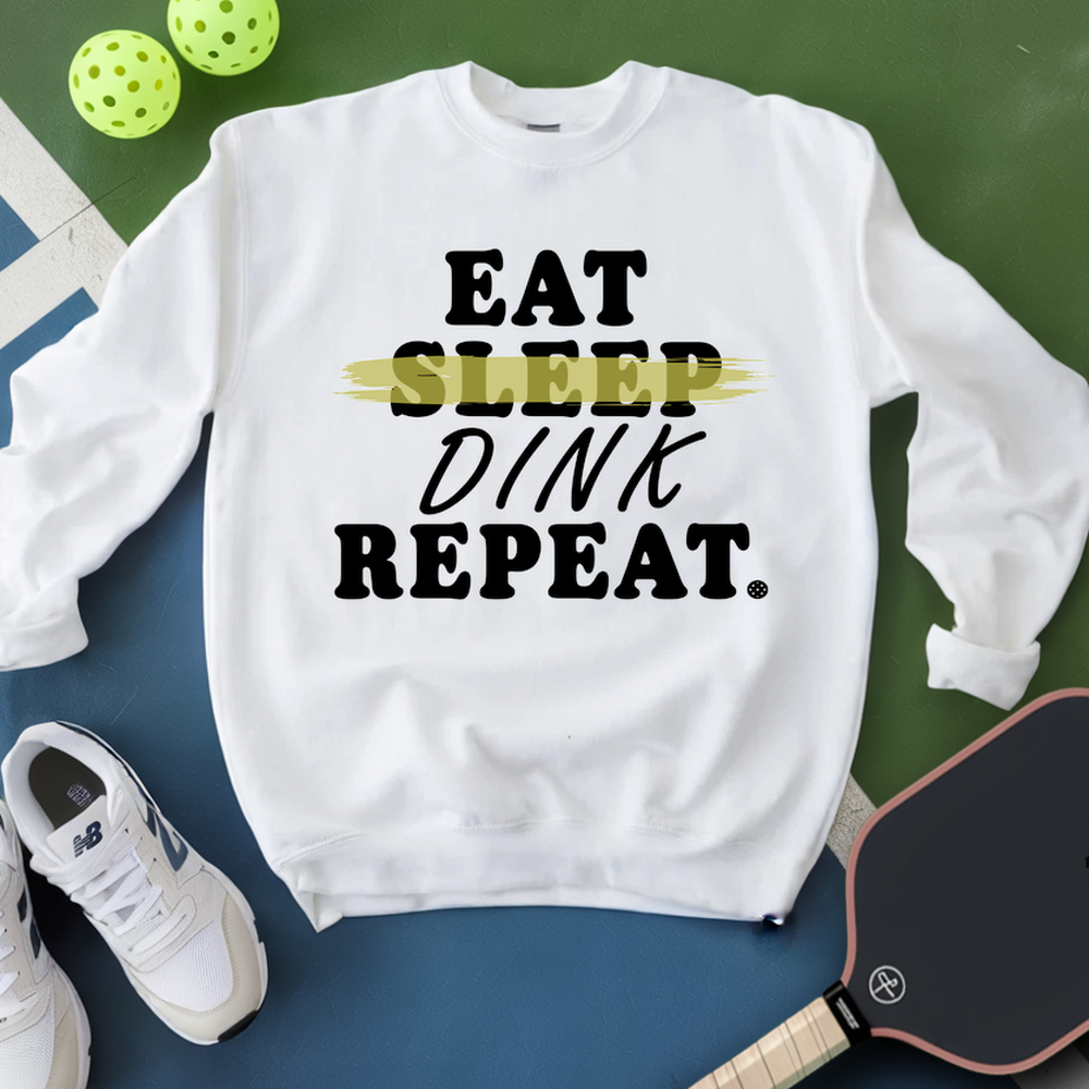 White sweatshirt with 'Eat Sleep Dink Repeat' text, next to a pickleball paddle and balls on a green and blue background.