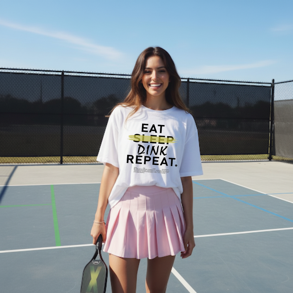 Woman on a tennis court wearing a white t-shirt with 'EAT SLEEP DINK REPEAT' text.