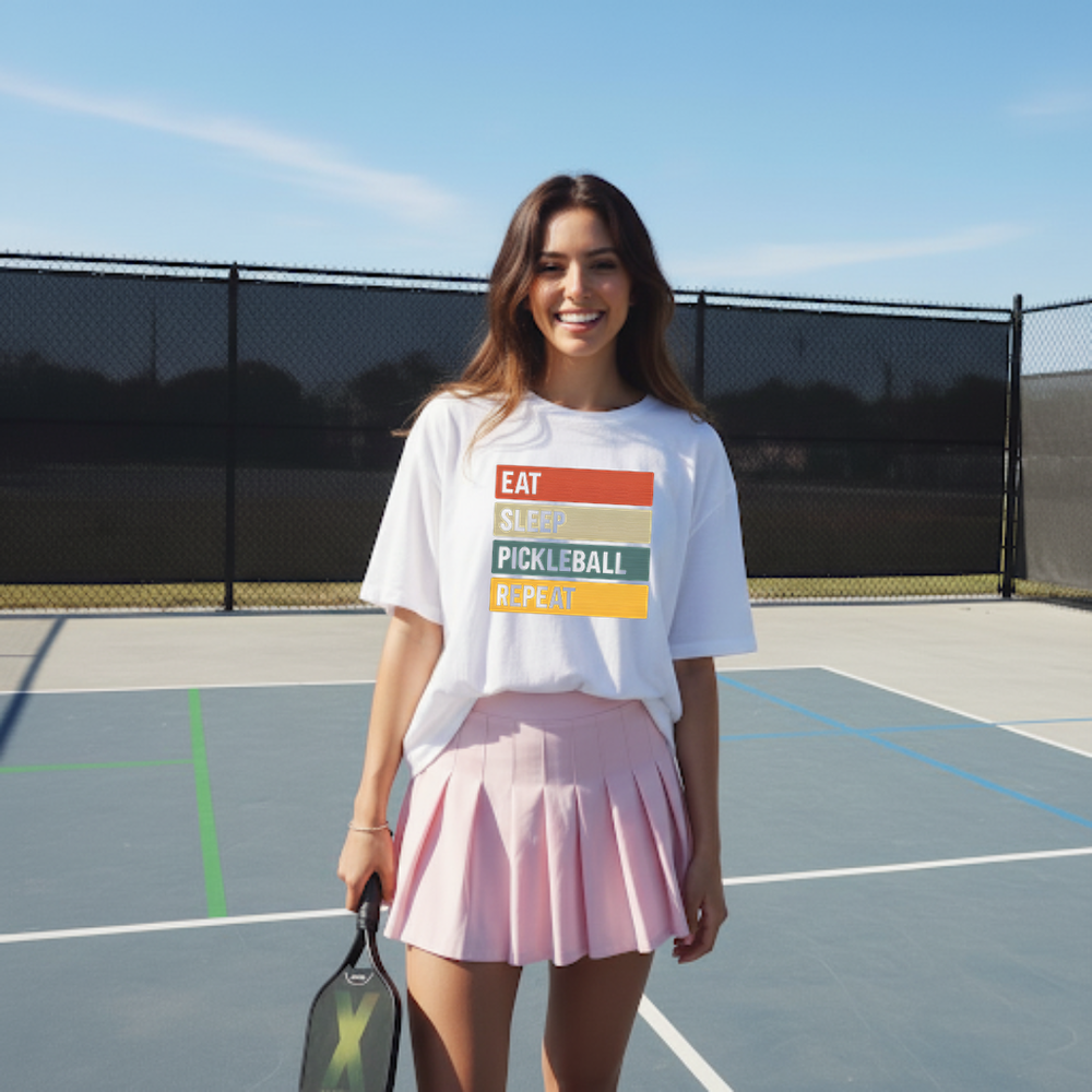 Woman on a pickleball court wearing a t-shirt with colorful text and a pink skirt.