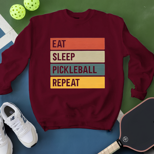Maroon sweatshirt with 'Eat Sleep Pickleball Repeat' text, pickleball paddles, and balls on a green and blue background