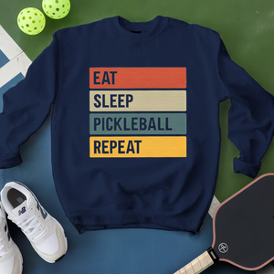 Navy blue sweatshirt with 'Eat Sleep Pickleball Repeat' text, surrounded by pickleballs and a paddle on a green surface.