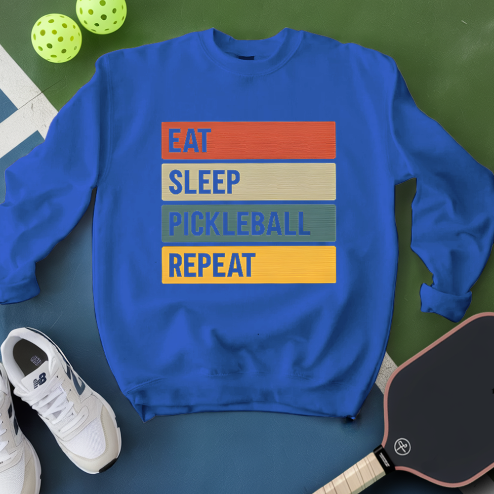 Blue sweatshirt with 'Eat Sleep Pickleball Repeat' text on a green surface with pickleballs and a paddle.