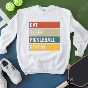 White sweatshirt with 'Eat Sleep Pickleball Repeat' text, pickleballs, and a paddle on a green and blue background
