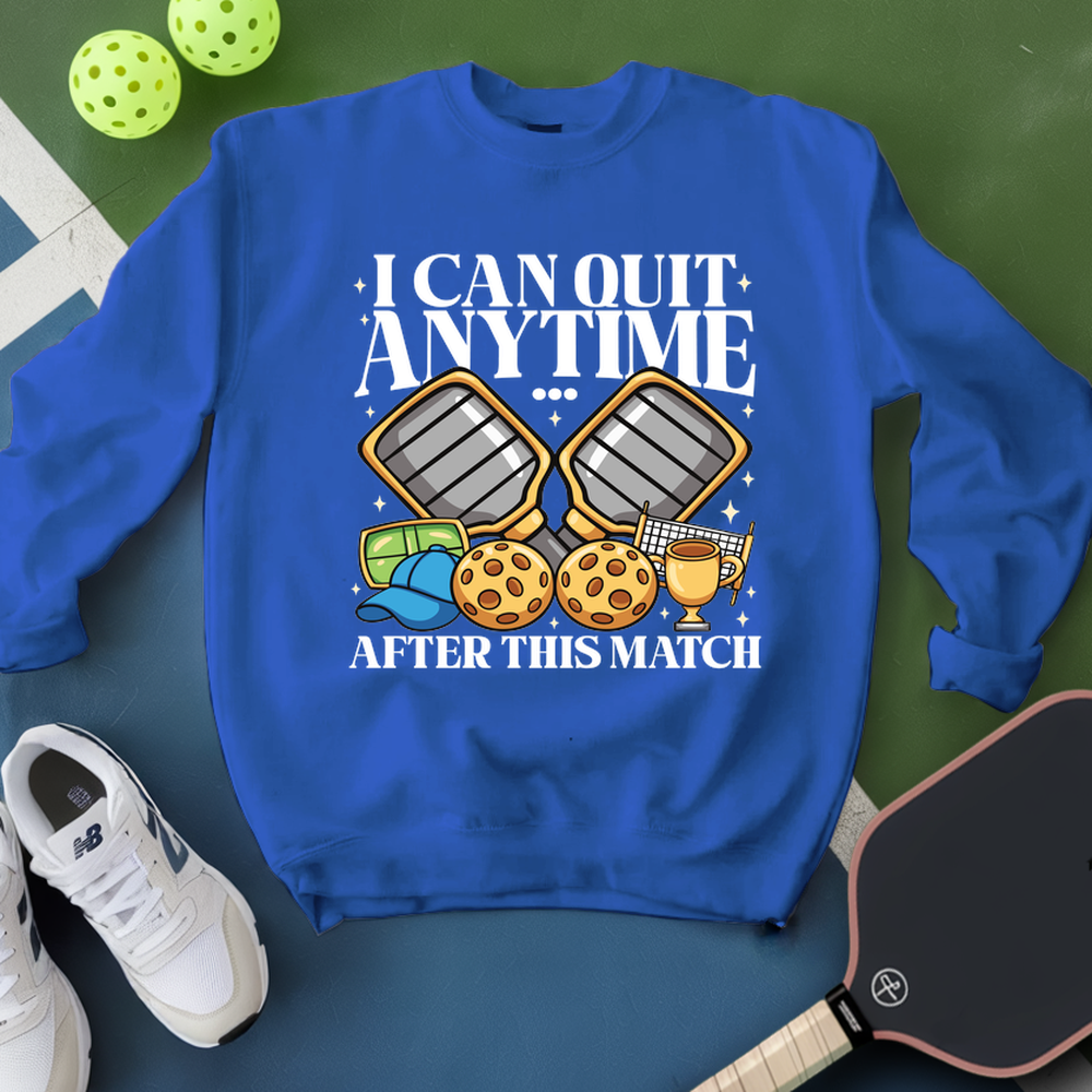 Blue sweatshirt with pickleball-themed design on a green surface with pickleballs and a paddle.