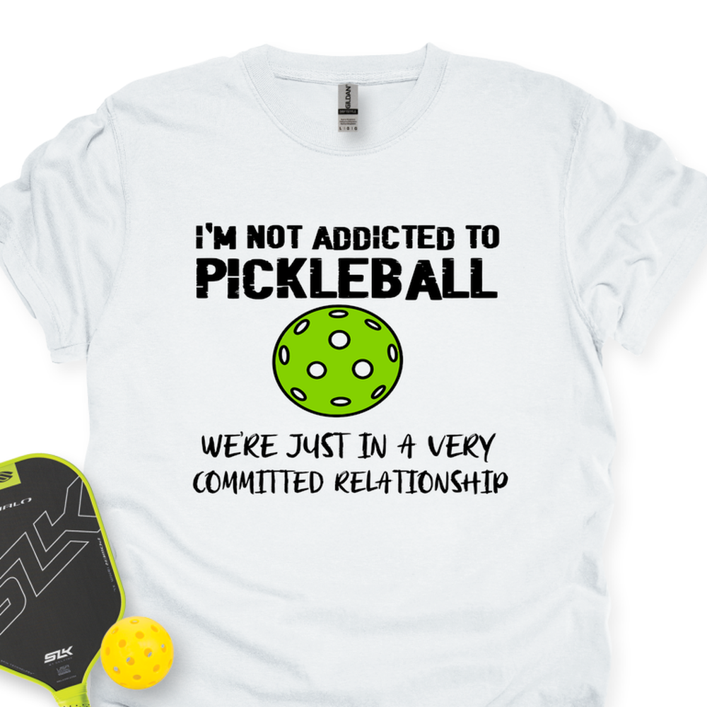 White t-shirt with pickleball-themed text and graphics, accompanied by a paddle and ball on a white background.