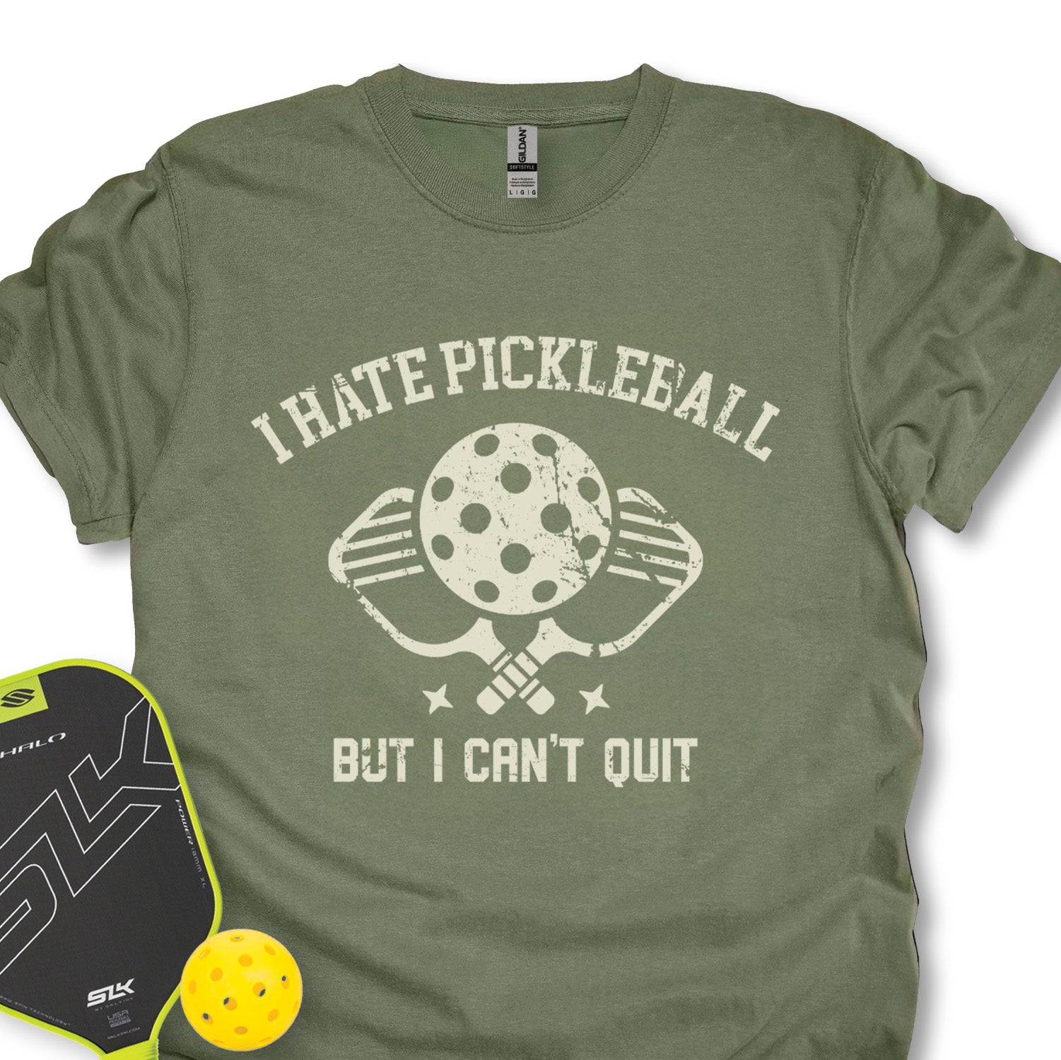 Green t-shirt with 'I Hate Pickleball But I Can't Quit' design, next to a pickleball paddle and ball on a white background.