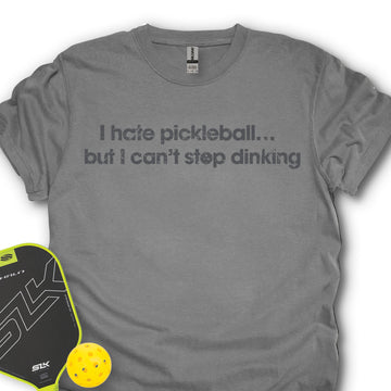 Gray t-shirt with humorous pickleball text, next to a pickleball paddle and ball on a white background