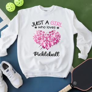 White sweatshirt with pink heart design and text on a green and blue surface with pickleball equipment.