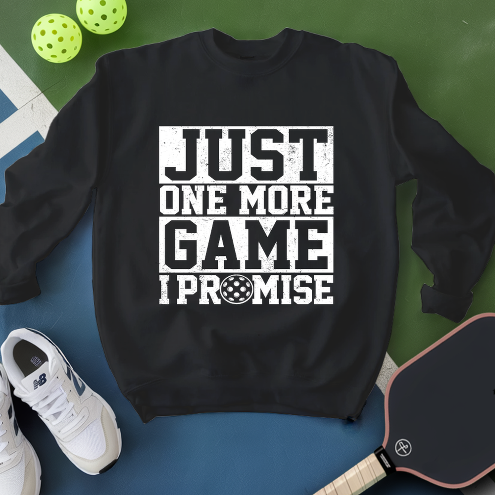 Black sweatshirt with 'Just One More Game I Promise' text, white sneakers, and pickleball equipment on a green and blue background.