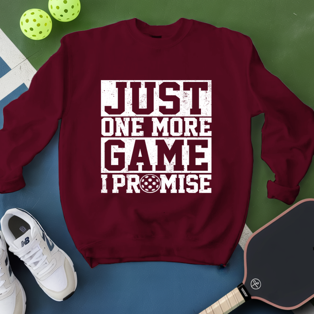 Maroon sweatshirt with 'Just One More Game I Promise' text, white sneakers, and pickleball equipment on a green and blue background.