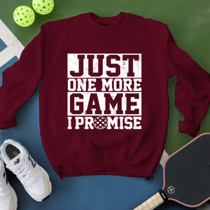 Maroon sweatshirt with 'Just One More Game I Promise' text, white sneakers, and pickleball equipment on a green and blue background.