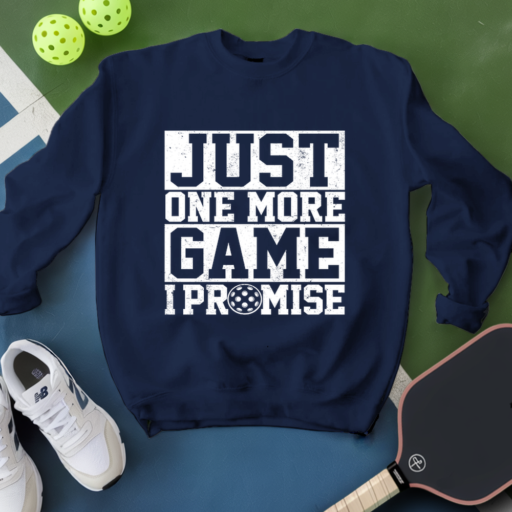 Navy blue sweatshirt with 'Just One More Game I Promise' text, white sneakers, and pickleball equipment on a green surface.