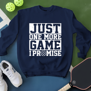 Navy blue sweatshirt with 'Just One More Game I Promise' text, white sneakers, and pickleball equipment on a green surface.