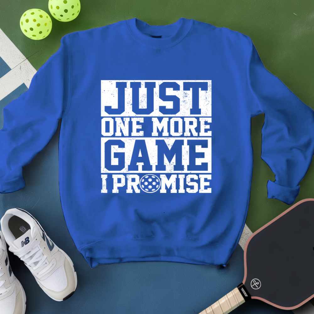 Blue sweatshirt with 'Just One More Game I Promise' text, white sneakers, and pickleball equipment on a green surface.
