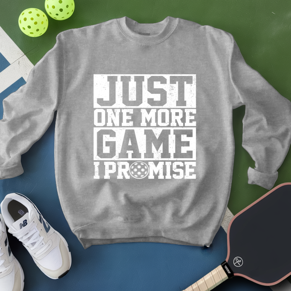 Gray sweatshirt with 'Just One More Game I Promise' text, white sneakers, and pickleball equipment on a green and blue background.