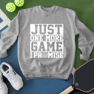 Gray sweatshirt with 'Just One More Game I Promise' text, white sneakers, and pickleball equipment on a green and blue background.