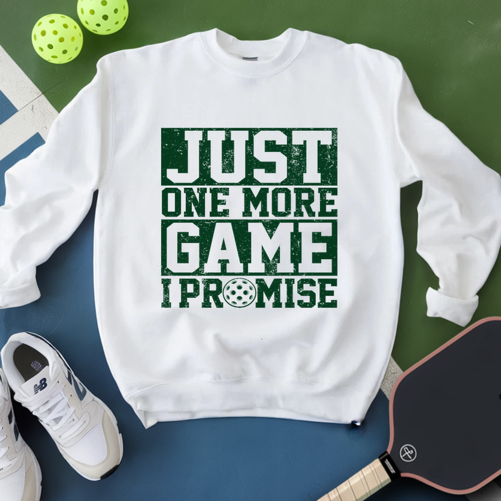 White sweatshirt with 'Just One More Game I Promise' text, next to pickleball equipment on a green and blue background.