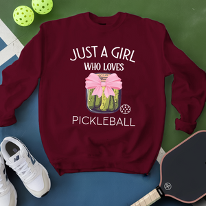 Maroon sweatshirt with pickleball design and text, placed on a green and blue surface with pickleball equipment.