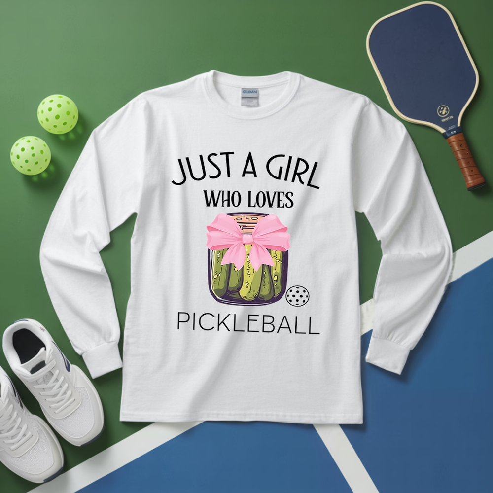 White long-sleeve shirt with pickleball design and text, placed on a green and blue surface with pickleball equipment.