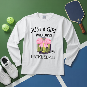White long-sleeve shirt with pickleball design and text, placed on a green and blue surface with pickleball equipment.
