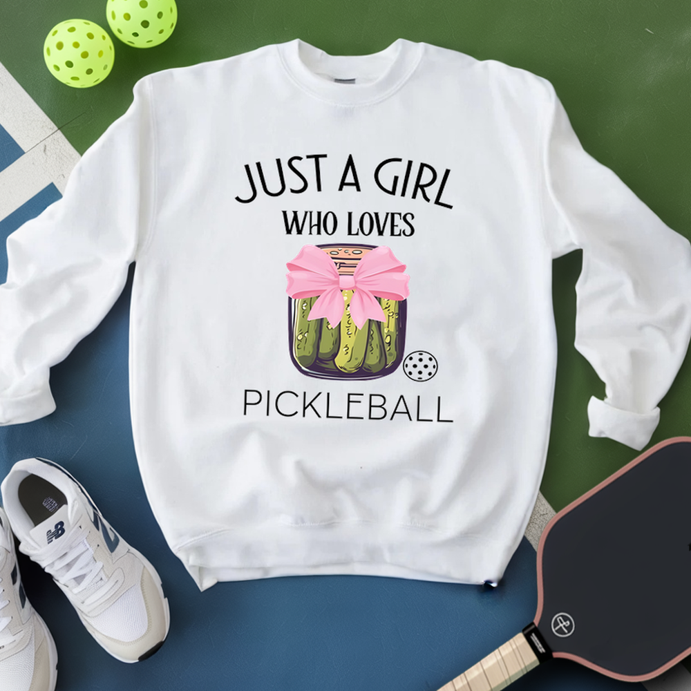 White sweatshirt with pickleball design and text, placed on a green and blue surface with pickleballs and a paddle.
