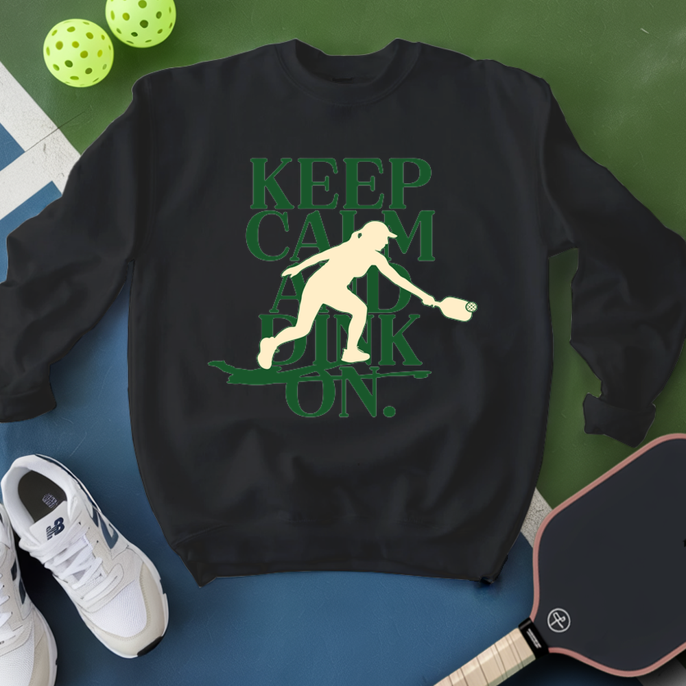 Black sweatshirt with green graphic and text, placed on a blue surface with pickleball equipment.