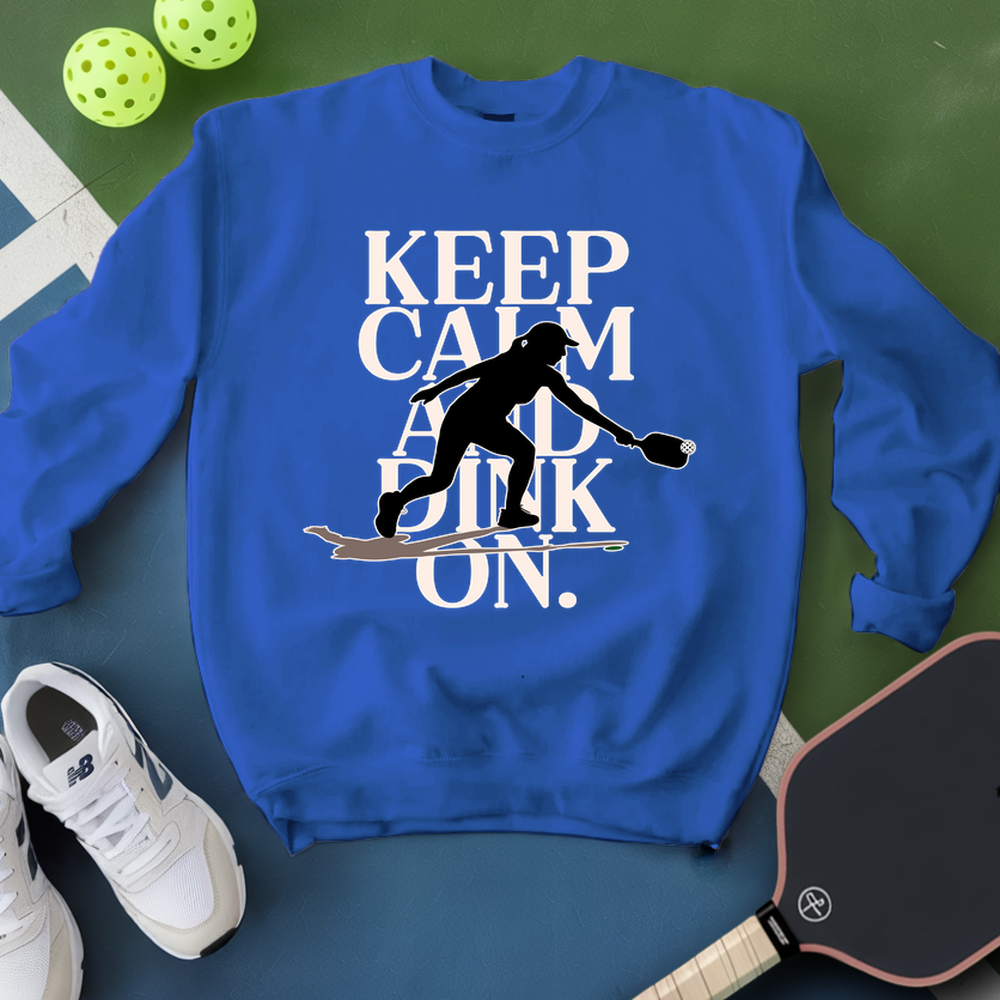 Blue sweatshirt with pickleball-themed design on a green surface with pickleballs and a paddle.