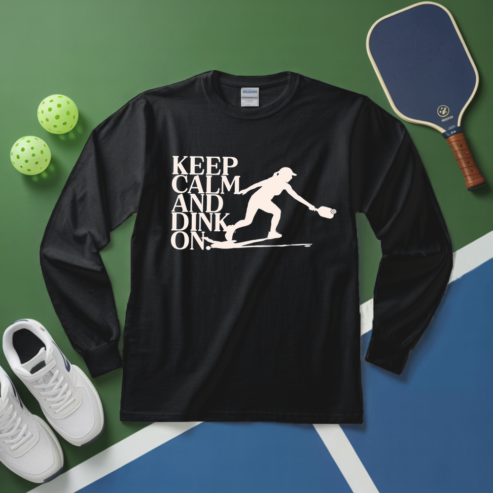Black long-sleeve shirt with 'Keep Calm and Dink On' graphic on a green and blue surface with pickleball equipment.