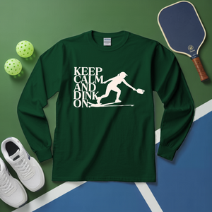 Green long-sleeve shirt with 'Keep Calm and Dink On' graphic on a green and blue surface with pickleball equipment.