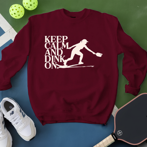 Maroon sweatshirt with 'Keep Calm and Dink On' graphic on a green and blue background with pickleball equipment.