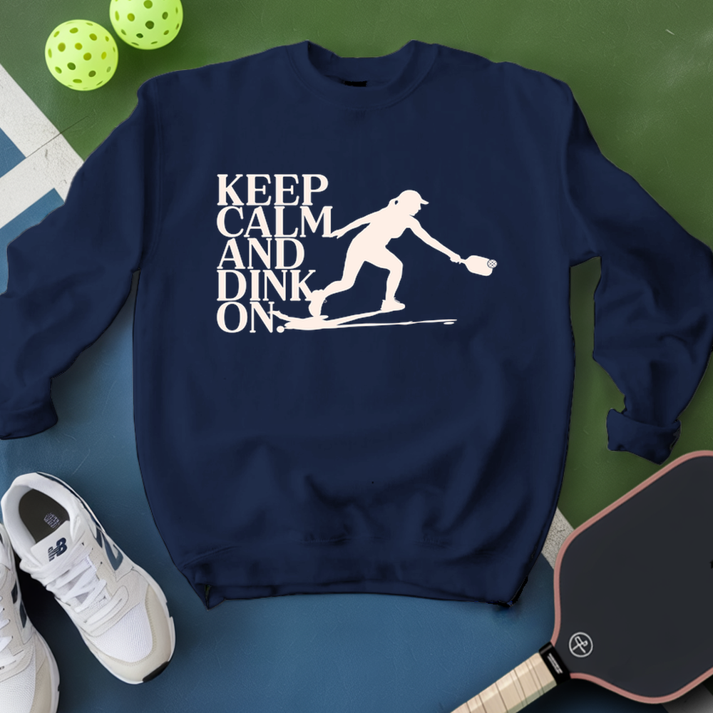 Navy blue sweatshirt with 'Keep Calm and Dink On' graphic on a green and blue surface with pickleball equipment.
