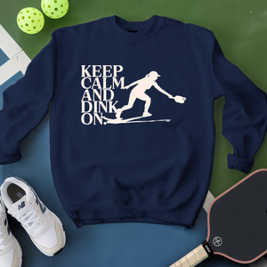 Navy blue sweatshirt with 'Keep Calm and Dink On' graphic on a green and blue surface with pickleball equipment.