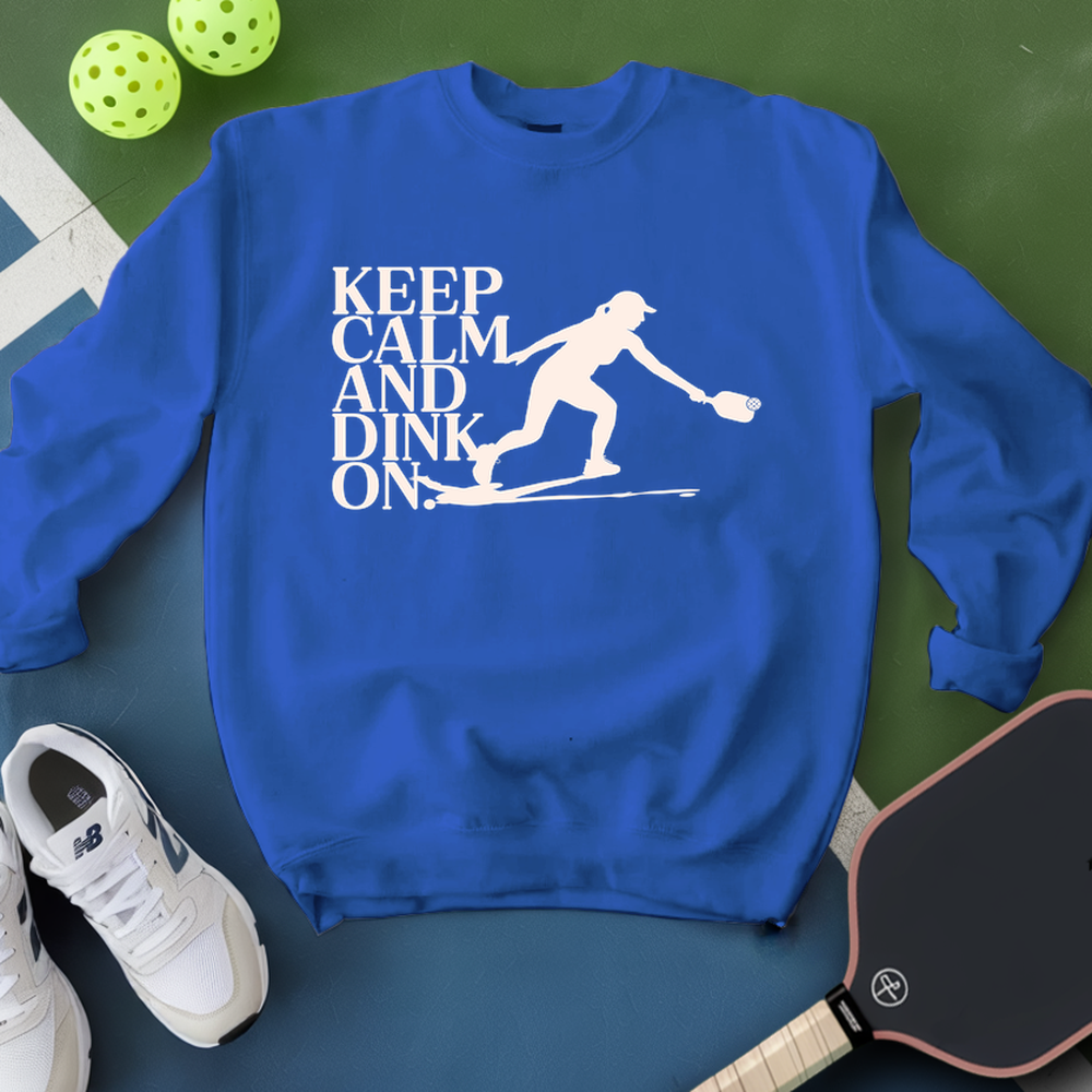 Blue sweatshirt with 'Keep Calm and Dink On' text and pickleball player graphic on a green surface with pickleballs and paddle.