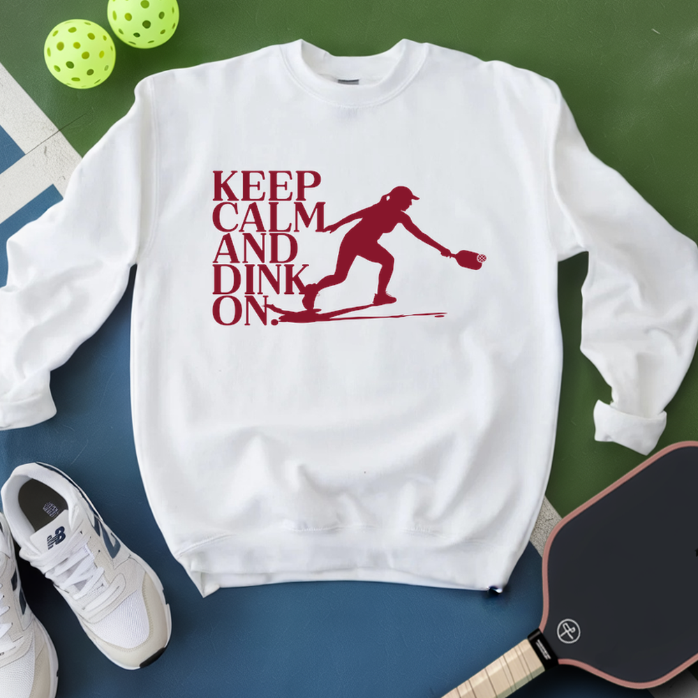 White sweatshirt with red graphic and text 'Keep Calm and Dink On' on a green and blue surface with pickleball equipment.