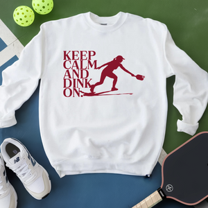 White sweatshirt with red graphic and text 'Keep Calm and Dink On' on a green and blue surface with pickleball equipment.